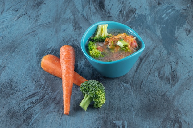 Zanahorias, brócoli y tazón de sopa de pollo, sobre la superficie azul.