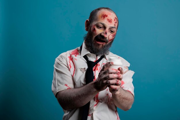 Zombi peligroso mirando una taza de café, queriendo tomar una bebida y siendo agresivo en el estudio. Monstruo malvado aterrador sosteniendo una taza y con heridas sangrientas, un diablo espeluznante apocalíptico.