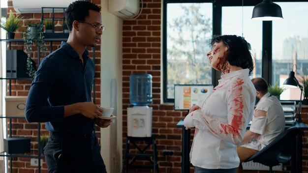 Zombie espeluznante hambriento hablando con un hombre de negocios mientras un compañero de trabajo muerto cubierto de sangre pasa caminando. Persona discutiendo con un monstruo no muerto malvado de aspecto extraño en la oficina mientras pasa un colega sin sentido.