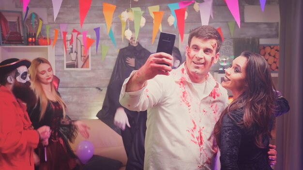Zombie peligroso y bruja espeluznante tomando un selfie en la fiesta de halloween en casa decorada