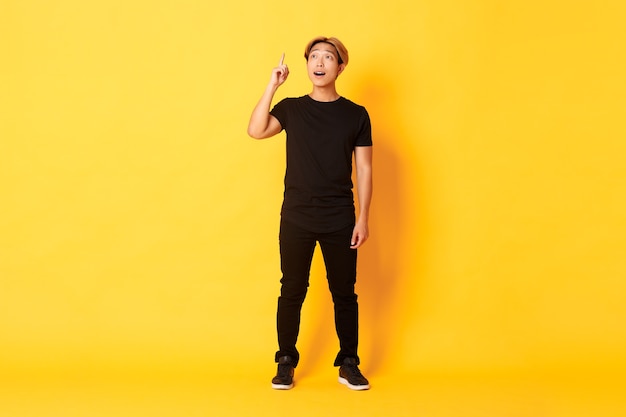 A figura intera di uomo asiatico bello premuroso, alzando il dito, avere una buona idea, in piedi muro giallo
