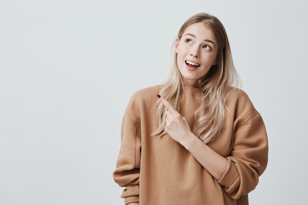 Abbastanza sorridente con gioia femminile con capelli biondi, guardando verso l'alto, indicando il dito indice, mostrando copia spazio