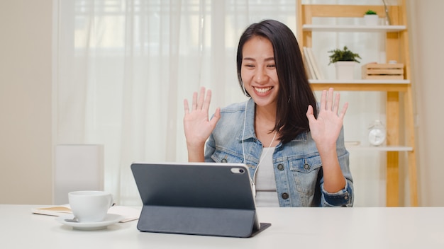 Abbigliamento casual da donna freelance che utilizza la videoconferenza di lavoro della compressa con il cliente in posto di lavoro in salone a casa. La giovane ragazza asiatica felice si rilassa la seduta sullo scrittorio fa il lavoro in Internet.