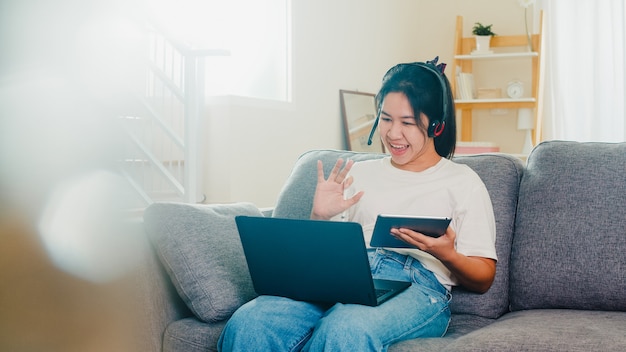 Abbigliamento casual freelance asiatico della donna di affari facendo uso della videoconferenza di lavoro del computer portatile con il cliente in posto di lavoro in salone da casa quando il distanziamento sociale resta a casa e tempo di quarantena di auto.