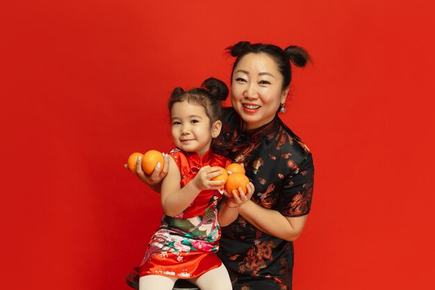 Abbracciare, sorridere, tenere in mano mandarini. . Ritratto asiatico della figlia e della madre sulla parete rossa in vestiti tradizionali. Celebrazione, emozioni umane, vacanze. Copyspace.