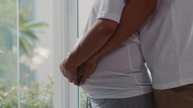 Abbraccio asiatico giovane delle coppie incinte e pancia della tenuta che parlano con il loro bambino. Mamma e papà sentirsi felici sorridenti pacifici mentre abbi cura del bambino, gravidanza vicino alla finestra nel salotto di casa.