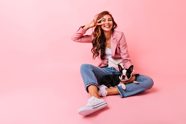 Accattivante giovane donna con i capelli lunghi in posa sul pavimento con il cane. Incredibile ragazza bruna seduta sul rosa con bulldog francese.