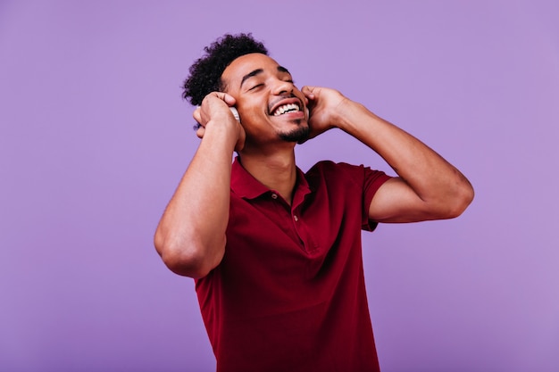 Accattivante uomo africano in maglietta rossa in posa con gli occhi chiusi e il sorriso timido. Foto dell'interno della musica d'ascolto del modello nero bello.