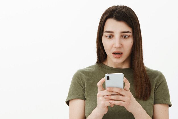 Accidenti, non indovina mai cosa è stato pubblicato online. Ritratto di ragazza colpita e senza parole utilizzando lo smartphone guardando stupito e sbalordito allo schermo del telefono cellulare con la bocca aperta dallo stupore oltre il muro grigio