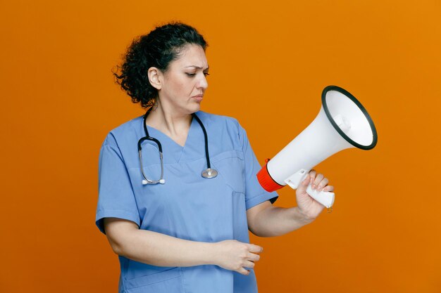 Accigliata dottoressa di mezza età che indossa uniforme e stetoscopio intorno al collo che tiene l'altoparlante guardandolo isolato su sfondo arancione