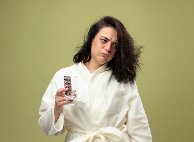 Accigliata giovane caucasica ragazza malata che indossa accappatoio holding pack di pillole mediche bicchiere d'acqua e tovagliolo guardando lato isolato su sfondo verde oliva