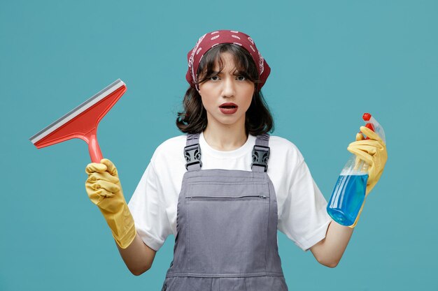 Accigliata giovane donna addetta alle pulizie che indossa bandana uniforme e guanti di gomma che mostrano tergicristallo e detergente guardando la fotocamera isolata su sfondo blu