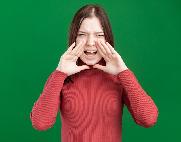 Accigliata giovane donna graziosa che tiene la mano vicino alla bocca guardando davanti chiamando ad alta voce qualcuno isolato sul muro verde