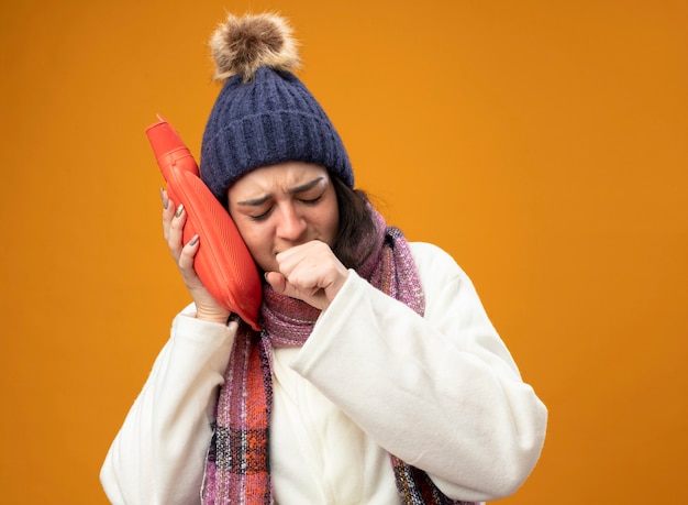 Accigliata giovane donna malata che indossa abito invernale cappello e sciarpa toccando la testa con la borsa dell'acqua calda tosse tenendo il pugno vicino alla bocca con gli occhi chiusi isolato sul muro arancione