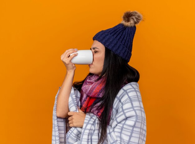 Accigliata giovane donna malata che indossa cappello invernale e sciarpa avvolta in un plaid in piedi in vista di profilo che beve tazza di tè con gli occhi chiusi isolati sul muro arancione