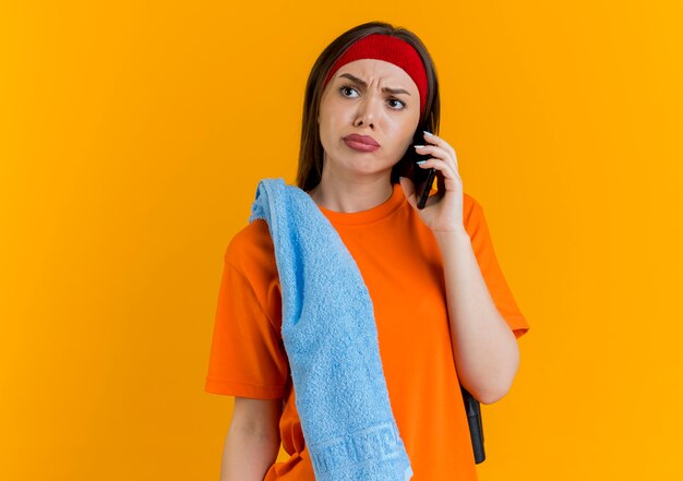 Accigliata giovane donna sportiva che indossa fascia e braccialetti con corda per saltare e asciugamano sulle spalle parlando al telefono guardando a lato