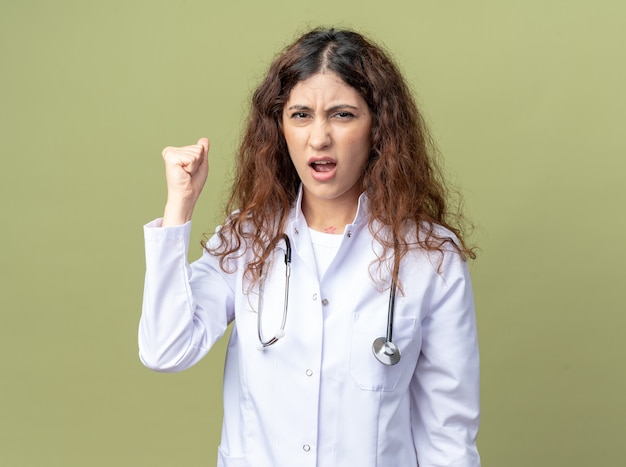 Accigliata giovane dottoressa che indossa una tunica medica e uno stetoscopio guardando la parte anteriore facendo un gesto forte isolato sulla parete verde oliva