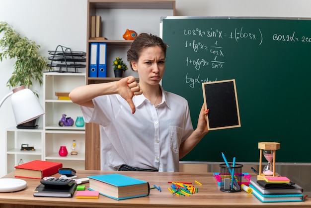 Accigliata giovane insegnante di matematica femminile bionda seduta alla scrivania con strumenti scolastici che tengono mini lavagna guardando la telecamera che mostra il pollice verso il basso in classe in