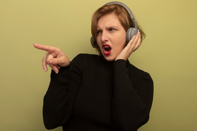 Accigliata giovane ragazza bionda che indossa le cuffie mettendo la mano su di loro guardando e puntando a lato