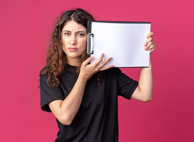 Accigliata giovane ragazza carina che tiene appunti toccando la testa con essa isolata sul muro cremisi