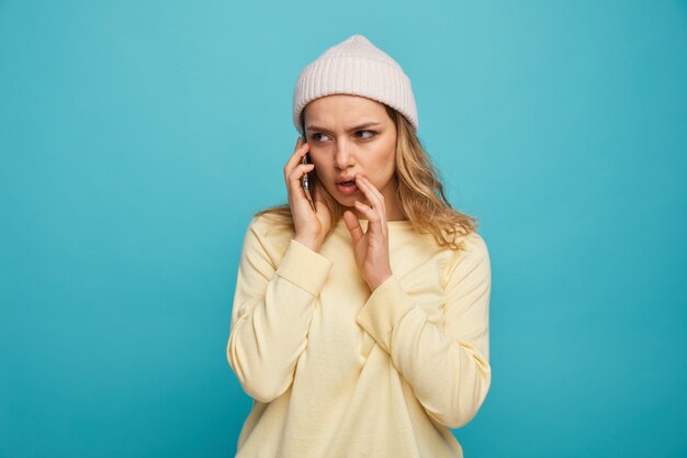 Accigliata giovane ragazza che indossa un cappello invernale parlando al telefono tenendo la mano vicino alla bocca sussurrando guardando a lato