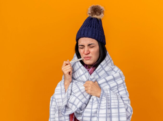 Accigliata giovane ragazza malata caucasica che indossa cappello invernale e sciarpa avvolta in plaid tenendo il termometro in bocca afferrando plaid con gli occhi chiusi isolato su sfondo arancione con spazio di copia