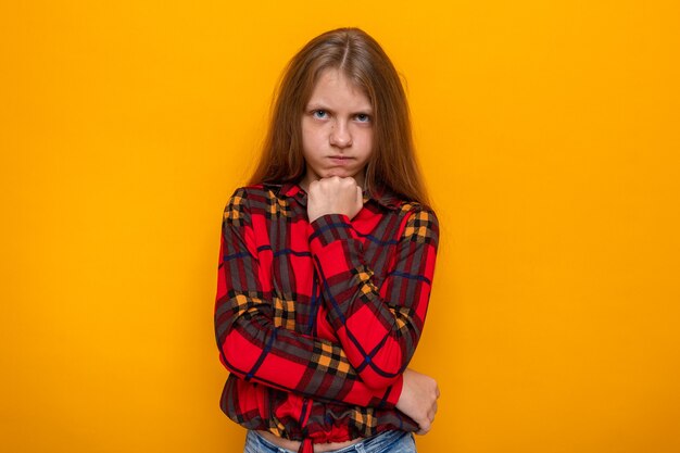 Accigliata mettendo la mano sotto il mento bella bambina che indossa una camicia rossa