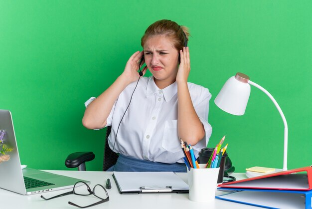 Accigliata ragazza bionda del call center che indossa l'auricolare seduto alla scrivania con strumenti di lavoro che tiene le mani sull'auricolare guardando il laptop