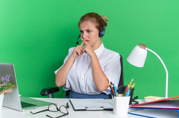 Accigliata ragazza bionda del call center che indossa le cuffie seduto alla scrivania con strumenti di lavoro che guarda il laptop senza fare gesti