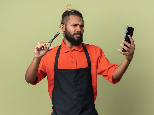 Accigliato giovane barbiere afroamericano che indossa un'uniforme con forbici e telefono cellulare guardando il telefono