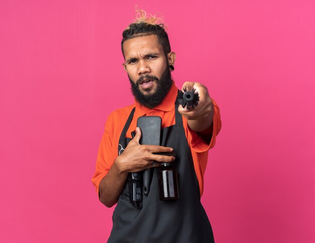 Accigliato giovane barbiere maschio afroamericano che indossa un'uniforme che tiene gli strumenti del barbiere guardando e puntando la telecamera con il pettine