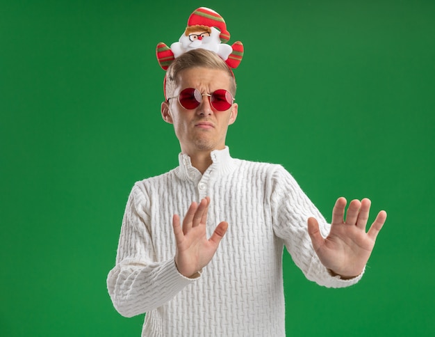 Accigliato giovane bel ragazzo che indossa la fascia di Babbo Natale con gli occhiali guardando il lato facendo gesto di rifiuto isolato sulla parete verde con lo spazio della copia