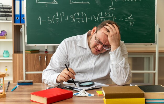 accigliato giovane insegnante con gli occhiali seduto alla scrivania con materiale scolastico in classe tenendo la matita tenendo la mano sulla testa guardando in basso