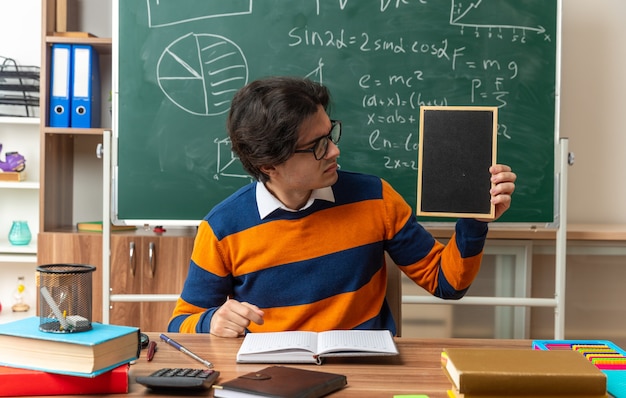 accigliato giovane insegnante di geometria con gli occhiali seduto alla scrivania con materiale scolastico in aula che mostra una mini lavagna a guardarla