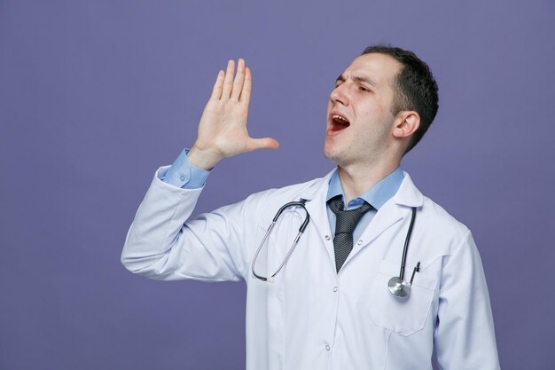 Accigliato giovane medico maschio che indossa accappatoio medico e stetoscopio intorno al collo guardando il lato tenendo la mano in aria chiamando qualcuno isolato su sfondo viola