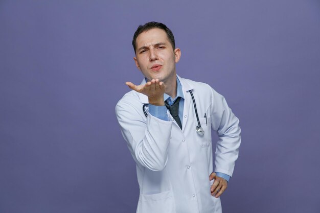 Accigliato giovane medico maschio che indossa accappatoio medico e stetoscopio intorno al collo tenendo la mano sulla vita guardando la telecamera che soffia bacio isolato su sfondo viola