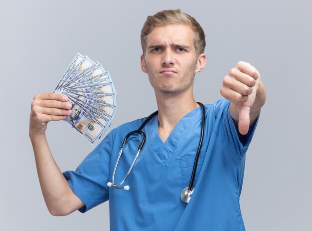 Accigliato giovane medico maschio che indossa l'uniforme del medico con lo stetoscopio che tiene contanti che mostra il pollice giù isolato sul muro bianco