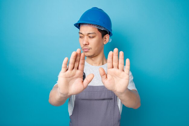 Accigliato giovane operaio edile che indossa il casco di sicurezza e uniforme guardando verso il basso facendo gesto di rifiuto