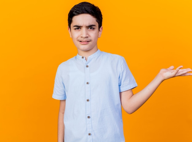 Accigliato giovane ragazzo caucasico che guarda l'obbiettivo che mostra la mano vuota isolata su priorità bassa arancione con lo spazio della copia