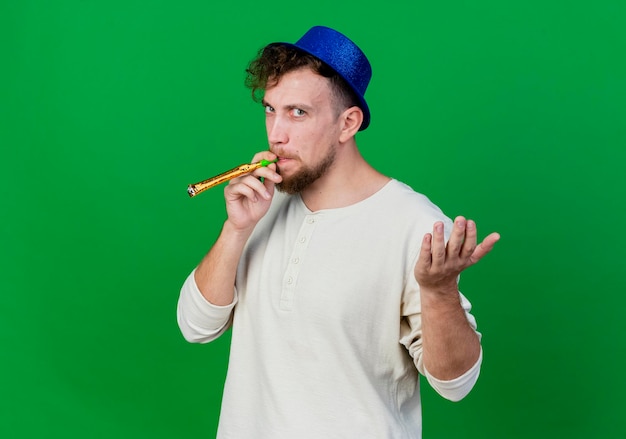 Accigliato giovane ragazzo slavo bello del partito che indossa il cappello del partito che soffia il ventilatore del partito che guarda davanti che mostra la mano vuota isolata sulla parete verde con lo spazio della copia