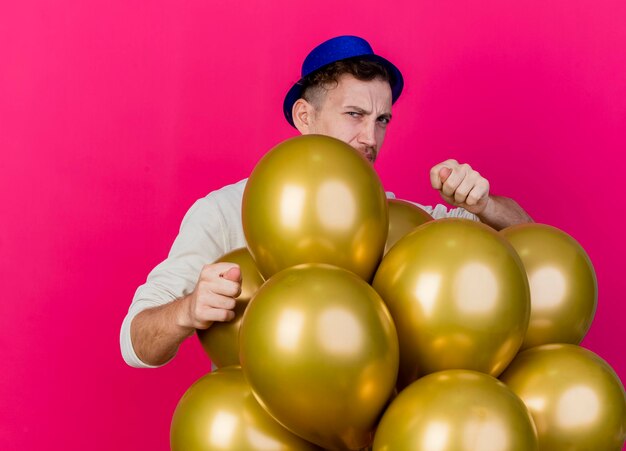 Accigliato giovane ragazzo slavo bello partito che indossa il cappello del partito in piedi dietro i palloncini guardando davanti facendo segno di fico isolato sul muro rosa