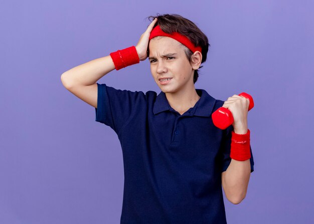 Accigliato giovane ragazzo sportivo bello che indossa la fascia e braccialetti con bretelle dentali che tengono il manubrio tenendo la mano sulla testa guardando il lato isolato sul muro viola