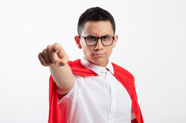 Accigliato giovane supereroe ragazzo in mantello rosso con gli occhiali guardando e indicando la fotocamera isolata su sfondo bianco con spazio di copia