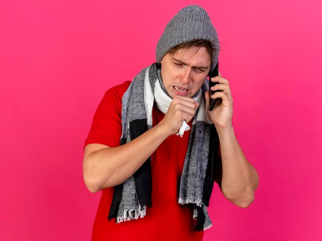 Accigliato giovane uomo malato bello biondo che indossa cappello invernale e sciarpa parlando al telefono tenendo il tovagliolo tenendo la mano vicino alla bocca tosse guardando in basso isolato sul muro rosa con lo spazio della copia