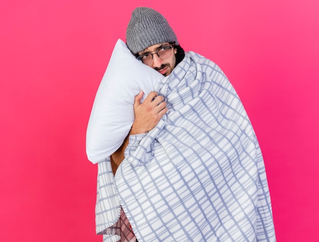 Accigliato giovane uomo malato con gli occhiali inverno cappello e sciarpa avvolto in plaid tenendo il cuscino toccando il viso con esso guardando il lato isolato sul muro rosa