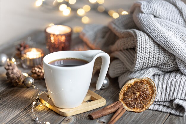 Accogliente composizione invernale domestica con una tazza di tè su uno sfondo sfocato con candele accese e luci bokeh ed elementi a maglia.