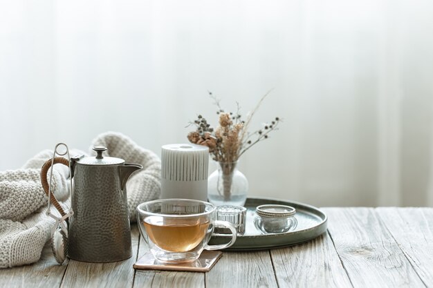 Accogliente natura morta con tazza di tè in vetro, candele ed elemento a maglia su sfondo sfocato spazio copia.
