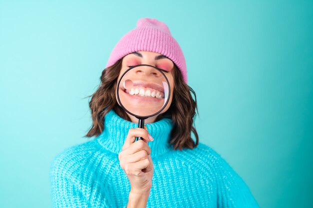 Accogliente ritratto di una giovane donna con un maglione blu lavorato a maglia e un cappello rosa con un trucco luminoso che tiene in mano una lente d'ingrandimento, scherzando, divertendosi