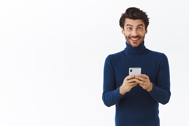 Acconciatura alla moda ragazzo sorridente allegro in maglione a collo alto con fotocamera sguardo smartphone soddisfatto e affascinato ricevere grandi notizie tramite messaggio in app prodotto ordinato con grande sconto