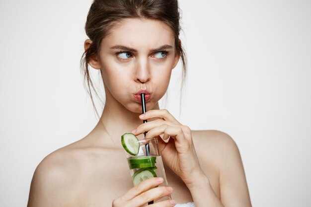 Acqua potabile dispiaciuta della ragazza con le fette del cetriolo sopra fondo bianco. Cosmetologia e spa di bellezza. Trattamento facciale.